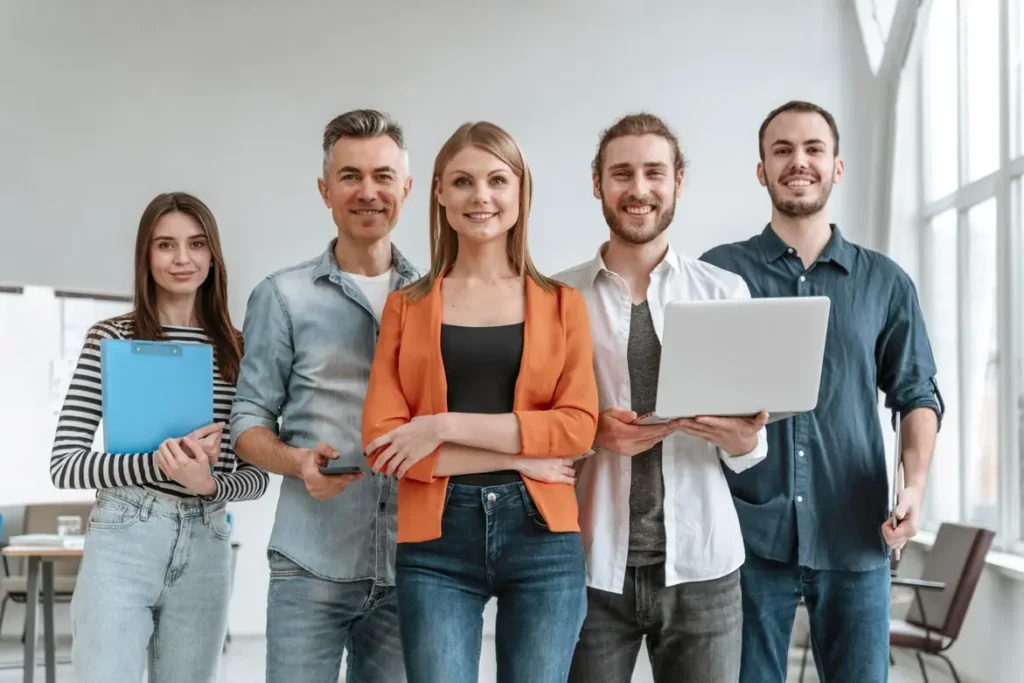 A team of five diverse and smiling professionals, representing the DIDITRA careers, stands confidently in an office environment, equipped with technology and documents, ready to deliver innovative solutions.